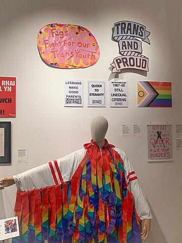 Signs showing solidarity with Trans people above an LGBTQ+ flag and a mannequin wearing a jacket with the pride rainbow colours hanging off it in strips.
